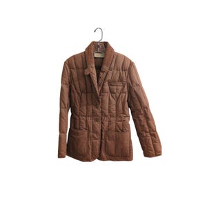 May include: A brown quilted jacket featuring a notched collar, a button-front closure, and two large patch pockets on the lower front. The garment has long sleeves and a vertical stitched pattern throughout the fabric, hanging on a clear hanger.