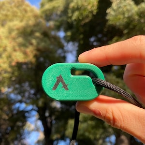 May include: A green, oval-shaped tag with a black 'A' logo, attached to a black cord. The tag is held up against a blurred background of green trees and a blue sky.