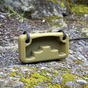 May include: Olive green rectangular object with a stepped interior design and two black cords attached. The object is resting on a mossy, rocky surface, suggesting an outdoor setting. The design appears to be 3D printed.