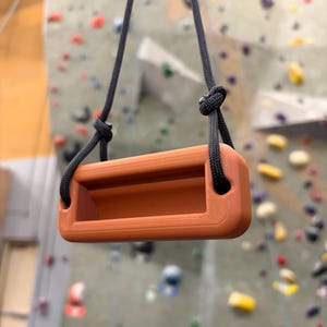 May include: A terracotta-colored rectangular climbing hangboard with a hollow center, suspended by a black braided rope that is knotted on each side. The training tool features a smooth, matte finish and rounded edges, designed for grip strength.