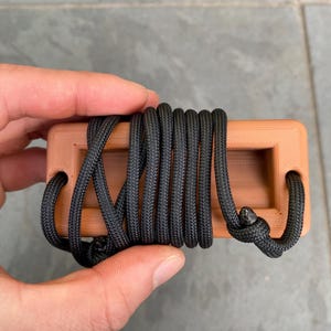 May include: A rectangular, light brown object with a black rope wrapped around it. The object has two holes on the sides and is held by a hand. The rope is knotted on one end.