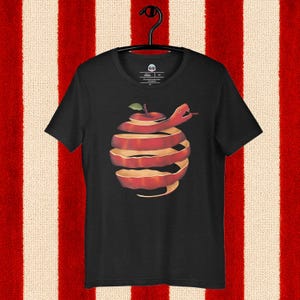 Könnte beinhalten: Schwarzes T-Shirt mit einem Motiv eines geschälten roten Apfels und einer roten Schlange. Das Design ist auf dem T-Shirt zentriert. Das T-Shirt hängt an einem schwarzen Kleiderbügel. Der Hintergrund hat rote und weiße Streifen.