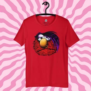 Puede incluir: Camiseta roja con un personaje de dibujos animados. El personaje tiene pelaje rojo, una nariz amarilla y pelo morado y negro. Tiene un piercing y una expresión melancólica. La camiseta está colgada de una percha negra sobre un fondo de rayas rosas y blancas.