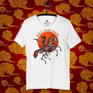 Op de afbeelding: Wit T-shirt met een afbeelding van een skeletcowboy die een skelethorse rijdt voor een rode zon. De tekst "Hell Yeah Haw!" staat rond de zon.