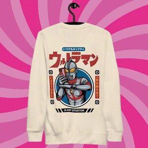 May include: A cream coloured sweatshirt featuring a graphic of a superhero character holding a smartphone. The graphic includes the text "ウルトラマン" (Ultraman) and "いつでもキャプチャ" (Capture anytime). The text "CAPTURE THE MOMENT IN ANY SITUATION" is also included in the graphic.