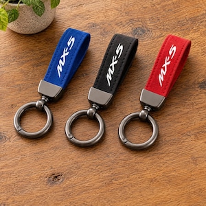 May include: Three keychains featuring a loop of blue, black, and red fabric with the white text MX-5 printed on each. Each keychain has a dark metallic connector and a circular metal ring attached to the bottom of the fabric loop.