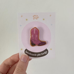 May include: A pink cowboy boot pin badge attached to a white display card. The boot features a darker pink toe and heel with white star accents. The card displays the text emy studio at the top and handmade pin badge in a black oval at the bottom.