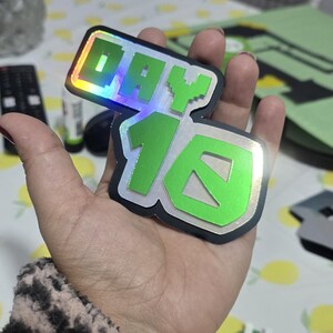 May include: Two stickers with the text "DAY 16" in a pixelated font. The stickers have a green and silver color scheme with a holographic effect. The stickers are held in a hand.