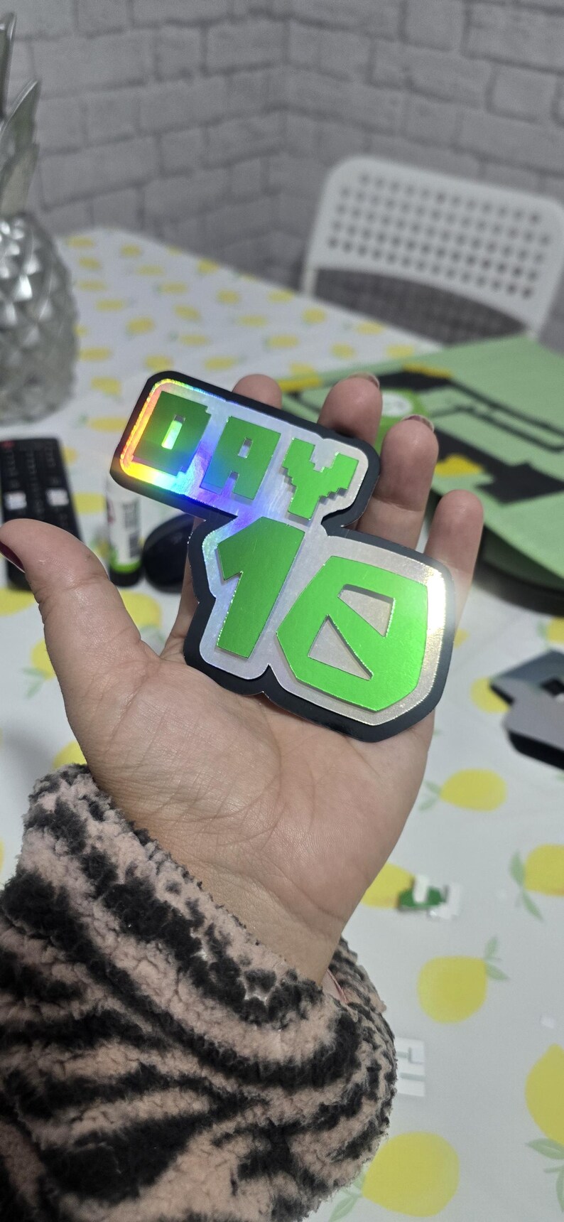 May include: Two stickers with the text "DAY 16" in a pixelated font. The stickers have a green and silver color scheme with a holographic effect. The stickers are held in a hand.