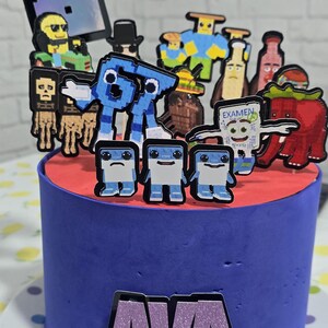 May include: A blue cake with a red top, decorated with pixelated character toppers. The cake has the name "AVA" on the front. The cake is on a white plate with colorful polka dots.