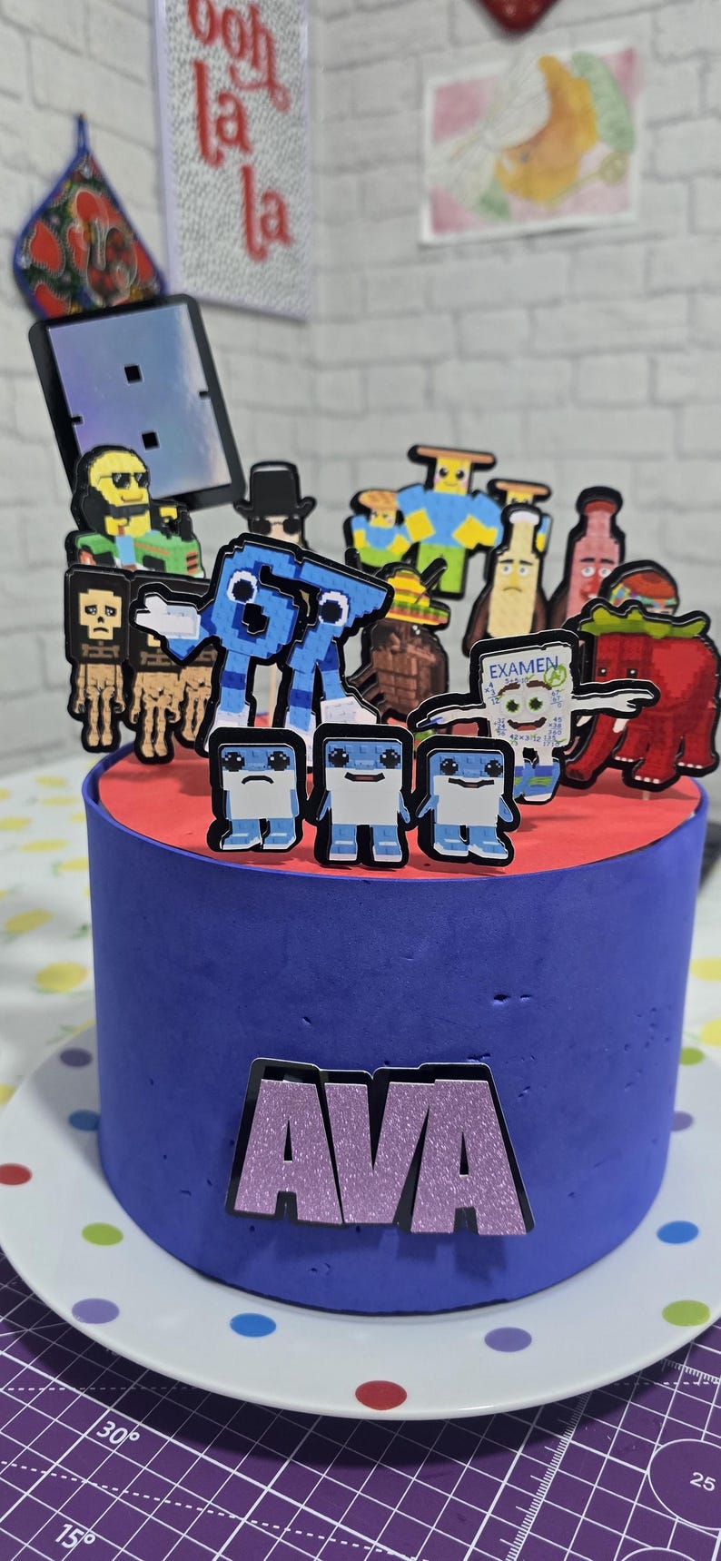 May include: A blue cake with a red top, decorated with pixelated character toppers. The cake has the name "AVA" on the front. The cake is on a white plate with colorful polka dots.
