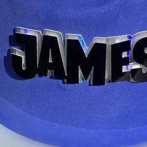May include: A blue frosted cake with the name "JAMES" in silver and black lettering. The cake sits on a white plate with a floral design. The cake is round and appears to be freshly made.