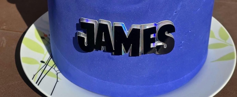 May include: A blue frosted cake with the name "JAMES" in silver and black lettering. The cake sits on a white plate with a floral design. The cake is round and appears to be freshly made.
