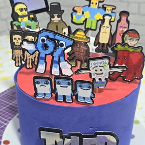 May include: A blue frosted cake with a red top, decorated with pixelated character toppers. The cake has the name "TYLER" on the side. The toppers include various figures, some holding signs, and a tablet-shaped decoration.
