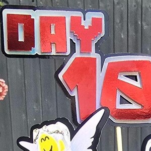 May include: Red and white Day 16 cake topper with a pixelated "Y" in "DAY". The numbers and letters have a shiny, reflective surface. The background is a blurred outdoor scene.