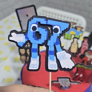 May include: A blue pixelated letter R cake topper with a black outline and white accents. The topper is attached to a wooden stick and is placed on a red and blue cake. The cake is decorated with other pixelated characters.