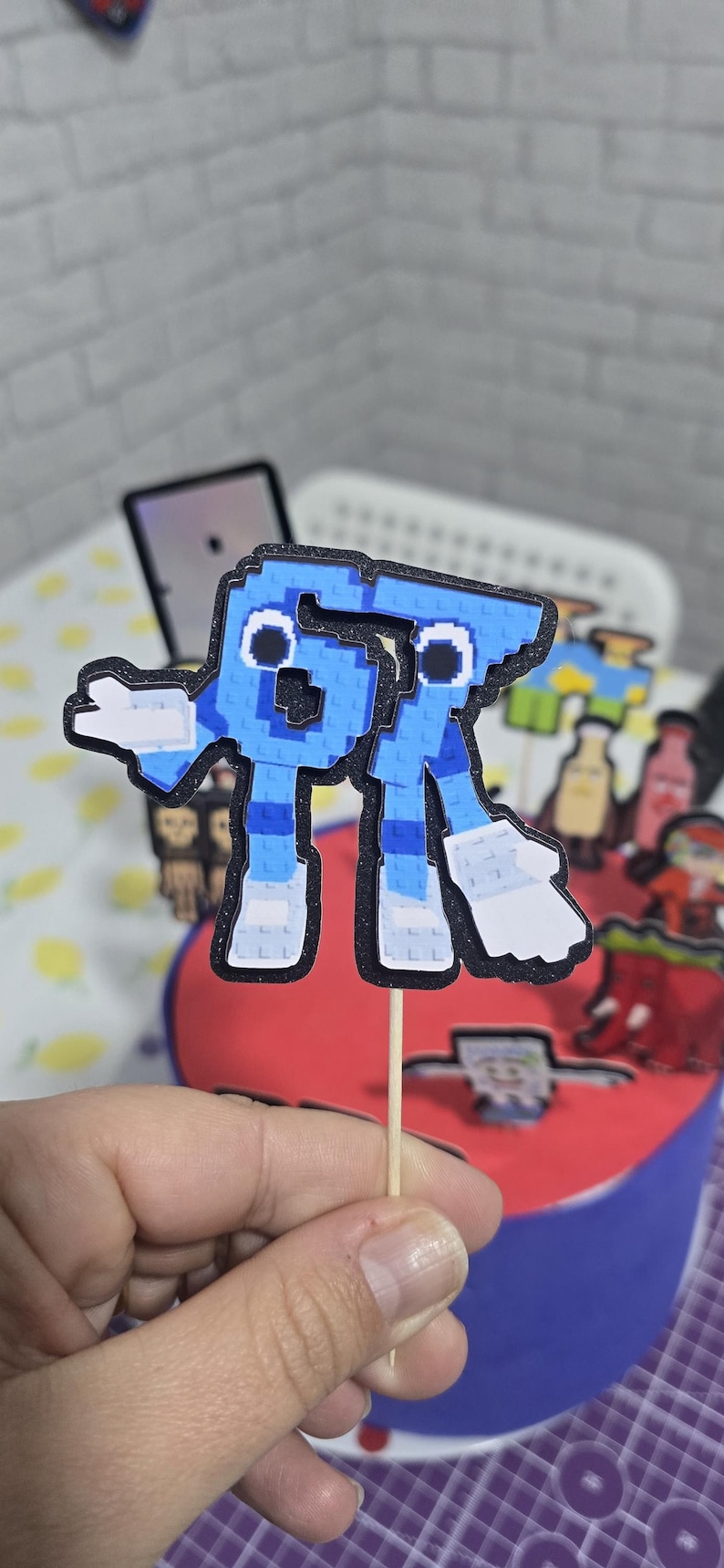 May include: A blue pixelated letter R cake topper with a black outline and white accents. The topper is attached to a wooden stick and is placed on a red and blue cake. The cake is decorated with other pixelated characters.