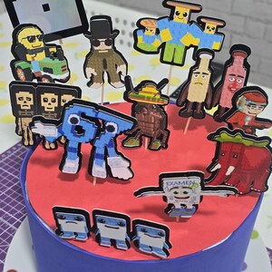 May include: A blue frosted cake with a red top, decorated with pixelated character toppers. The cake is adorned with the name "Tyler" in silver letters. The cake sits on a plate with colorful polka dots.