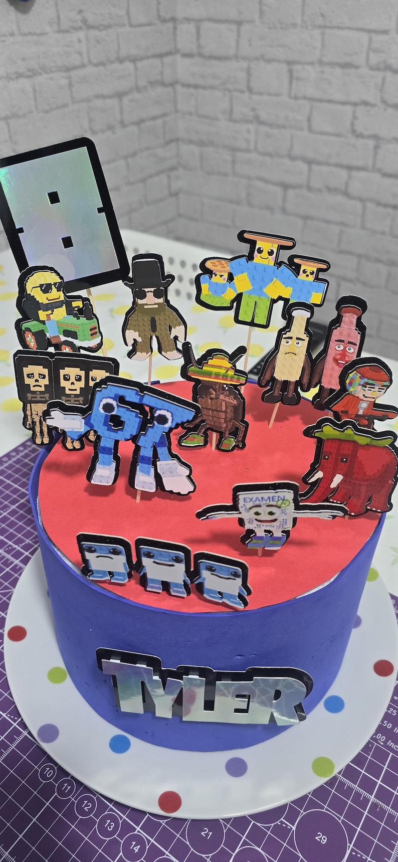May include: A blue frosted cake with a red top, decorated with pixelated character toppers. The cake is adorned with the name "Tyler" in silver letters. The cake sits on a plate with colorful polka dots.