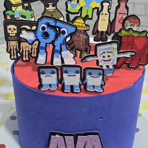 May include: A round cake with blue frosting and a red top layer. The cake is decorated with various pixelated character toppers. The name "AVA" is displayed on the front of the cake in glittery letters.