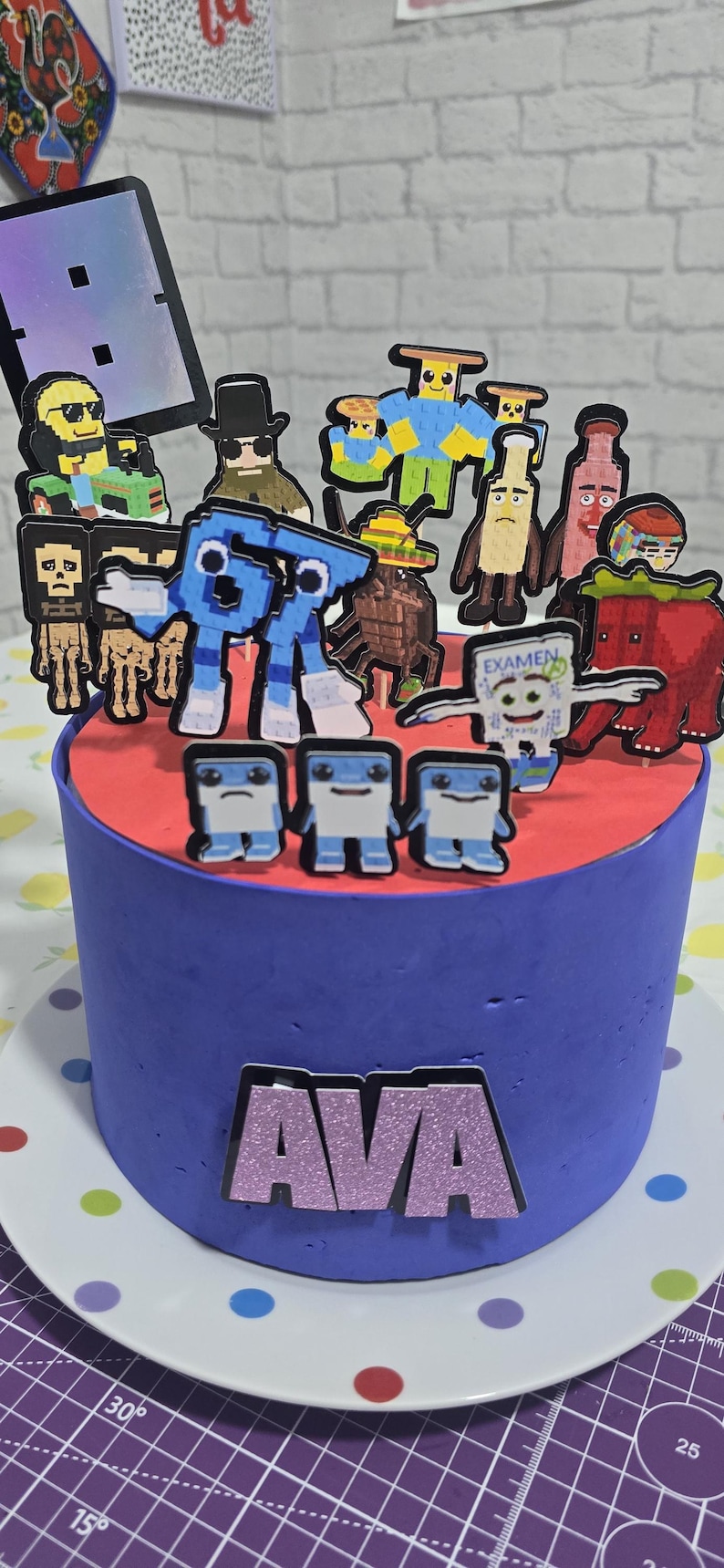 May include: A round cake with blue frosting and a red top layer. The cake is decorated with various pixelated character toppers. The name "AVA" is displayed on the front of the cake in glittery letters.