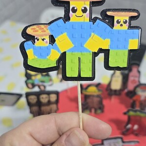 May include: A cupcake topper featuring three cartoon characters with yellow heads, blue shirts, and green pants. The characters are on a wooden stick, with a black outline. The background is blurred.
