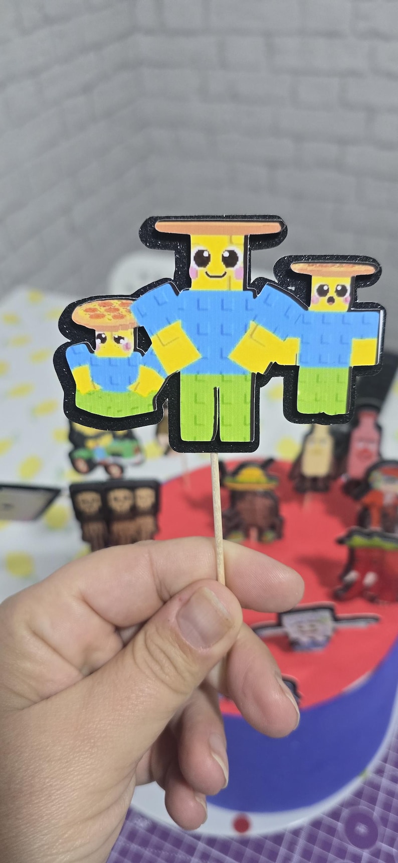 May include: A cupcake topper featuring three cartoon characters with yellow heads, blue shirts, and green pants. The characters are on a wooden stick, with a black outline. The background is blurred.