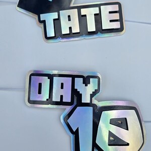 May include: Holographic stickers with black outlines and text. One sticker features a black square with a white square cutout. Another says "TATE" in capital letters. The third sticker says "DAY 16".
