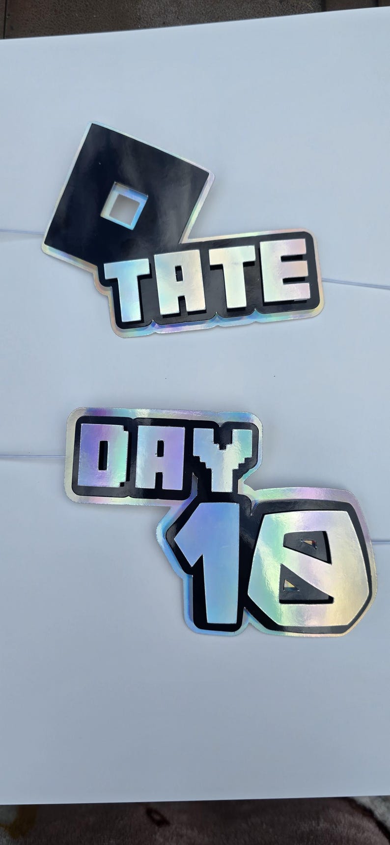 May include: Holographic stickers with black outlines and text. One sticker features a black square with a white square cutout. Another says "TATE" in capital letters. The third sticker says "DAY 16".