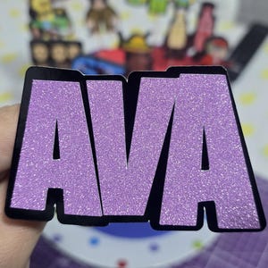 May include: A glittery purple sticker with the name "AVA" in bold, capital letters. The letters are outlined in black. The sticker is held in a hand, with other items visible in the background.
