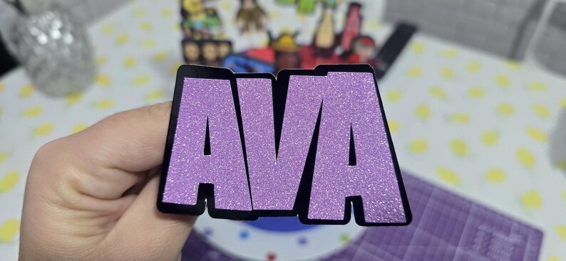 May include: A glittery purple sticker with the name "AVA" in bold, capital letters. The letters are outlined in black. The sticker is held in a hand, with other items visible in the background.