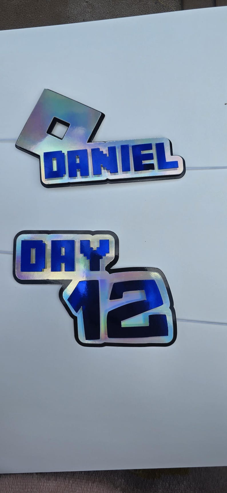 May include: Two-piece cake toppers with the name "DANIEL" and the text "DAY 12" in blue, outlined in black. The toppers have a holographic, iridescent finish and are set against a white background.