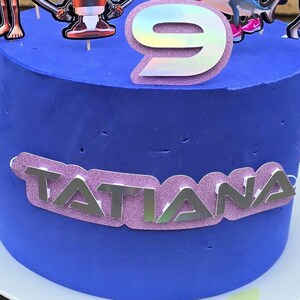 May include: A blue frosted birthday cake with a silver number 9 topper and character decorations. The name "Tatiana" is displayed in silver letters on a pink glitter banner around the cake.