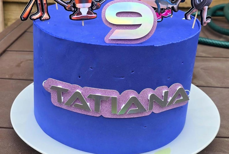 May include: A blue frosted birthday cake with a silver number 9 topper and character decorations. The name "Tatiana" is displayed in silver letters on a pink glitter banner around the cake.