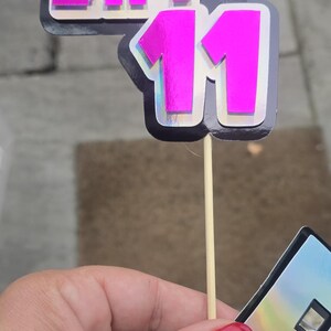 May include: A pink and silver Day 11 cake topper with a pixelated font. The number 11 is in a bold, shiny pink color. The topper is attached to a wooden stick.