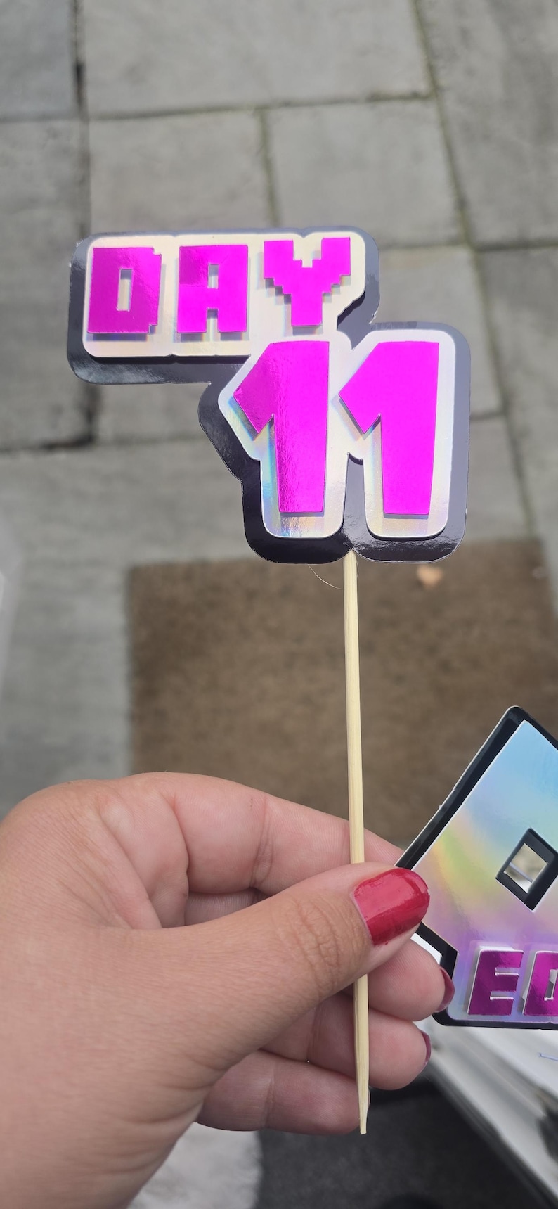 May include: A pink and silver Day 11 cake topper with a pixelated font. The number 11 is in a bold, shiny pink color. The topper is attached to a wooden stick.