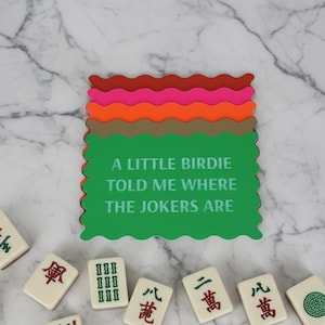 Może przedstawiać: Zielony podkładka pod kubek z napisem "A LITTLE BIRDIE TOLD ME WHERE THE JOKERS ARE" jest ułożony z innymi podkładkami w odcieniach czerwieni, r&oacute;żu, pomarańczy i brązu. Kafelki do mahjonga są rozrzucone.