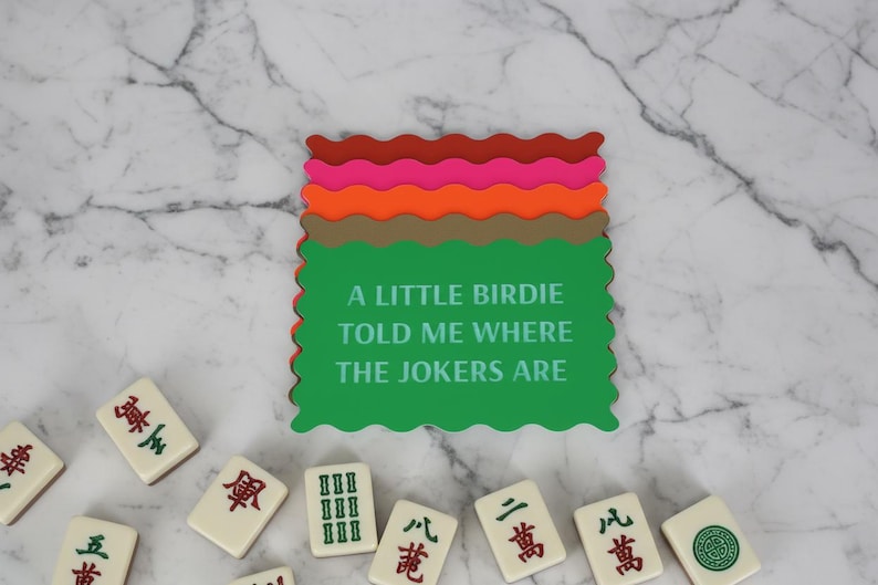 Może przedstawiać: Zielony podkładka pod kubek z napisem "A LITTLE BIRDIE TOLD ME WHERE THE JOKERS ARE" jest ułożony z innymi podkładkami w odcieniach czerwieni, r&oacute;żu, pomarańczy i brązu. Kafelki do mahjonga są rozrzucone.