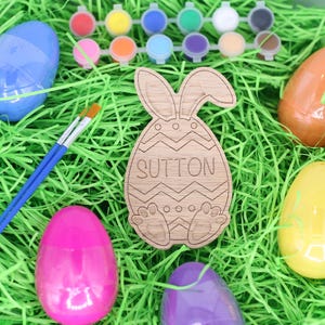 May include: An Easter-themed arrangement featuring a wooden bunny-shaped ornament with the name "SUTTON" engraved on it. The scene includes colorful plastic Easter eggs, paint pots, and paintbrushes, all set on a bed of green decorative grass.
