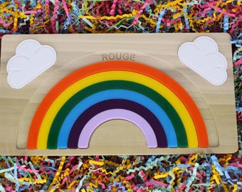 Könnte beinhalten: Holz-Regenbogen-Puzzle mit einem mehrfarbigen Bogen und zwei weißen Wolkenformen. Das Wort "ROUGE" ist auf der Holzbasis aufgedruckt. Das Puzzle befindet sich vor einem farbenfrohen Hintergrund aus geschreddertem Papier.