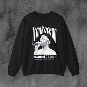 May include: Black sweatshirt with a white graphic print of Frank Ocean singing into a microphone. The text "frank ocean" is printed in white above the image and "channel ORANGE" is printed in white below the image.