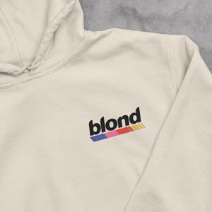May include: A beige hoodie with a black "blond" logo and a rainbow stripe underneath.