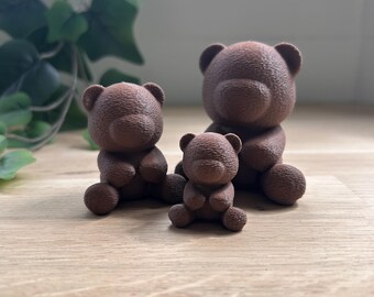Puede incluir: Tres figuras de osos de peluche marrones de diferentes tamaños están dispuestas sobre una superficie de madera. Los osos tienen un aspecto texturizado. Se ven hojas verdes en el fondo. El oso más pequeño está al frente.