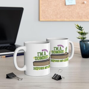 May include: Two white ceramic mugs with the text "Warning: I'm a Zombie Before My Coffee" in green and purple. The mugs feature a cartoon zombie rising from a coffin. The mugs are on a light-colored surface.