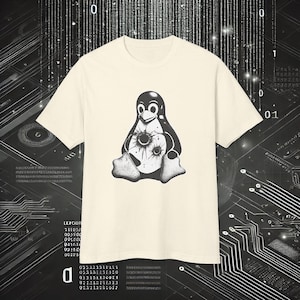 Può includere: T-shirt color crema con grafica in bianco e nero di Tux, la mascotte di Linux, con disegni di fori di proiettile. Il pinguino è seduto. La maglietta è a girocollo e maniche corte.