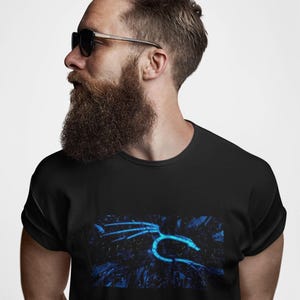 May include: Black t-shirt featuring a glowing blue graphic design. The design appears to be a stylized representation of a creature or symbol. The t-shirt is worn by a person with a beard and sunglasses.