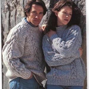 Könnte beinhalten: Zwei Personen tragen graue Strickpullover mit Zopfmuster. Der Mann trägt eine blaue Jeans und die Frau trägt eine blaue Jeans. Der Hintergrund ist eine Steinmauer.