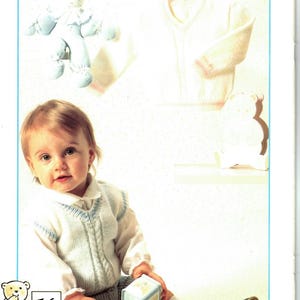 Può includere: Un bambino indossa un gilet e pantaloncini lavorati a maglia bianchi e azzurri, con una camicia bianca sotto. Il bambino tiene in mano un blocchetto blu e bianco. Un maglione bianco e un peluche sono sullo sfondo.