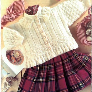 Può includere: Un cardigan in maglia bianca con colletto a volant e chiusura frontale con bottoni, abbinato a una gonna a pieghe a quadri rossi e neri. Il logo del marchio Patons è visibile in alto e un codice a barre con il testo Printed in Canada è in basso.