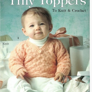May include: A baby wearing a peach-colored knit sweater with a textured pattern, white pants, and a white turtleneck. The image is a book cover titled "Tiny Toppers" with the text "To Knit & Crochet". The cover also includes the text "6 Designs By Marion Kelley".